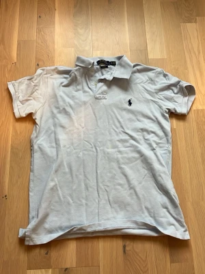 Vit pikétröja från Polo Ralph Lauren - Säljer en klassisk vit pikétröja från Polo Ralph Lauren i slim fit. Tröjan har korta ärmar, krage och två knappar framtill. Den är tillverkad i mjuk bomull och har den ikoniska broderade loggan på bröstet. Perfekt för en clean och stilren look.