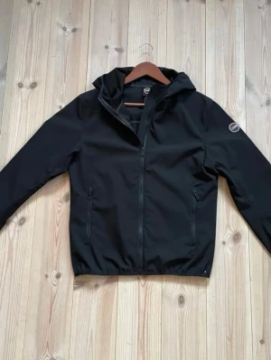 Colmar Vindjacka - Säljer en svart vindjacka från Colmar med huva och dragkedja framtill. Jackan har två fickor med dragkedja och en stilren design. 