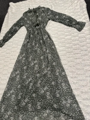 Grön blommig långklänning  - Säljer en långklänning i mörkgrönt med vitt blommigt mönster. Klänningen har långärmade ärmar, knytband med tofs vid halsen och lätt rynkad midja. Materialet är mjukt och tunt, perfekt för vår och sommar.