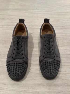 Loubs shoes  - Säljer ett par mörkgrå sneakers i mocka med coola svarta nitar på tån. Skorna har rund tå, platt sula och snörning. Insidan är beige och de har en stilren, urban vibe som sticker ut med detaljerna framtill. Använda i 2 veckor. Nytt skick.