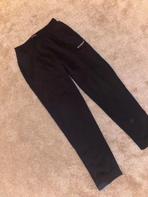 Svarta mjukisbyxor från McKenzie - Svarta mjukisbyxor från McKenzie med snörning i midjan och ribbade muddar vid bensluten. Byxorna har en enkel, clean look med logga tryckt på ena benet. Perfekta för chill eller träning. Materialet är mjukt och bekvämt, troligtvis bomull eller polyester.