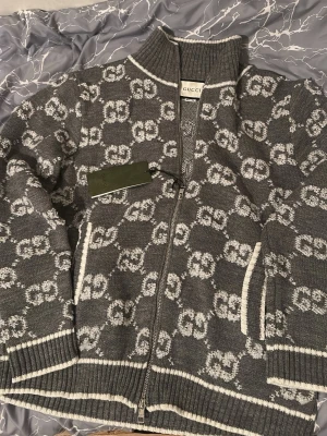 Grå stickad kofta från Gucci - Säljer en grå stickad kofta från Gucci med vit GG-mönster över hela plagget. Koftan har dragkedja framtill, ribbad krage och muddar samt vita detaljer vid fickor och kanter. Tillverkad i 100% ull, perfekt för kyliga dagar. Märke och storlek syns tydligt på etiketten.