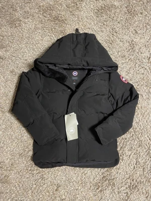 Svart dunjacka från Canada Goose M - Säljer en svart dunjacka från Canada Goose i storlek M. Jackan har huva, klassisk patch på ärmen och stängs med dragkedja och knappar. Perfekt för kalla dagar och har en clean, stilren look. Materialet är slitstarkt och jackan är vadderad för extra värme.