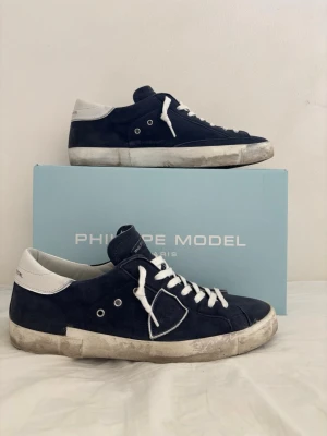 Philippe Model blå sneakers - Snygga sneakers från Philippe Model i mörkblå mocka med vita detaljer på hälen och vita snören. Skorna har en rund tå, platt sula och klassisk logga på sidan. Perfekta för dig som gillar stilrena och sportiga skor med en fräsch vibe. Låda och sko påse kommer med.