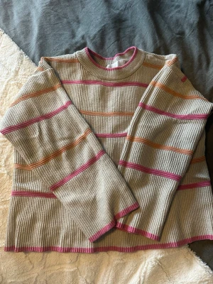 Randig ribbad tröja från SAMSA - Säljer en beige ribbad tröja från SAMSA Collection med långärmad modell. Tröjan har färgglada ränder i rosa och orange samt rund halsringning med rosa kant. Perfekt för dig som gillar retrovibbar och vill ha en mysig och snygg look.