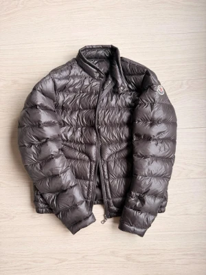 Mörkgrå dunjacka från Moncler - Säljer en mörkgrå dunjacka från Moncler i model Acorus med klassisk quiltad design och hög krage. Jackan har dragkedja framtill och Moncler-logga på ärmen. Materialet är glansigt och lätt, perfekt för kyliga dagar. Stilren look som funkar till allt. Jackan har små hål vid handlederna och så är även ena dragkedjan sönder man alla delar fås med för att laga den därav det billiga priset.
