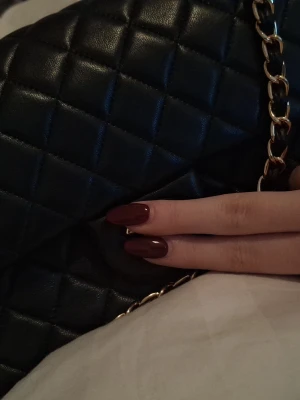 Svart quiltad axelväska med kedja - Säljer en svart axelväska med quiltad design och snygg guldfärgad kedja som axelrem. Väskan har en klassisk rektangulär form och är tillverkad i skinnimitation. Perfekt för dig som gillar stilrena detaljer och vill ha en väska som sticker ut.