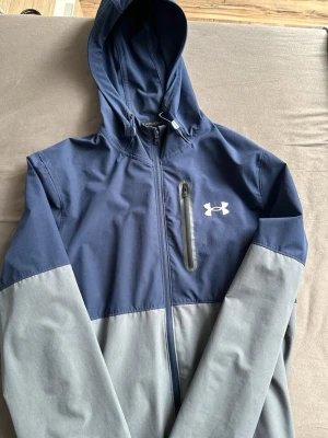  Vindjacka Under Armour - Säljer en snygg tvåfärgad vindjacka från Under Armour i marinblått och grått. Jackan har huva med dragsko, bröstficka med dragkedja och hel dragkedja framtill. Perfekt för blåsiga dagar och sportiga outfits.