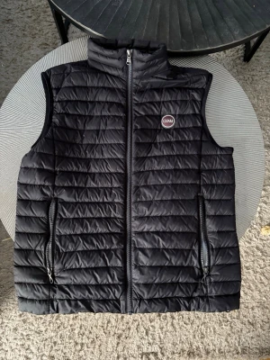 ÄKTA Svart dunväst från Colmar - Säljer en svart dunväst från Colmar med quiltad design och två praktiska fickor med dragkedja. Västen har en hög krage och logotyp på bröstet. Perfekt att slänga över en hoodie eller tröja när det är kyligt ute.