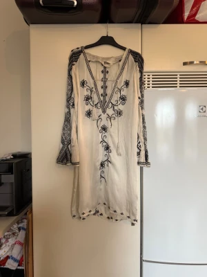 Vit och svart broderad kortklänning - Supersnygg vit kortklänning från Odd Molly med svart broderat blommönster och detaljer längs ärmar och hals. Klänningen har v-ringning med snörning och långa ärmar. Materialet är mjukt och känns som bomull, perfekt för sommarens alla tillfällen. Storlek 1 som enligt deras storleksguide motsvarar en storlek S. den ör fortfarande otroligt fin men vid broderier på klänningen kan det finnas lösa trådar. Mer använd. 