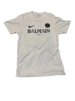Vit Balmain x PSG t-shirt Nike - Snygg vit t-shirt från Balmain x PSG i samarbete med Nike. Tryck med Balmain Paris och PSG-logga på bröstet, samt Nike swoosh. Klassisk rund hals och korta ärmar. Perfekt för dig som gillar streetwear och sportig stil.