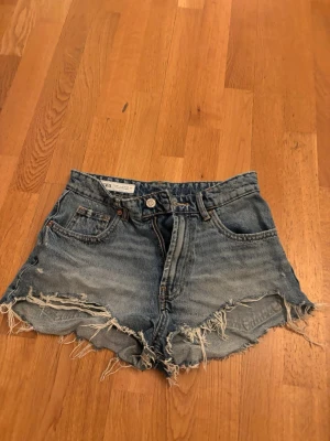 Blå jeansshorts från Zara - Säljer ett par blå jeansshorts från Zara med råa, fransiga kanter. Klassisk femficksmodell med knapp och dragkedja framtill.