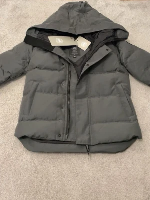 Canada Goose jacka  - Säljer en mörkgrå pufferjacka från Canada Goose med huva och snyggt svart logomärke på ärmen. Jackan har dragkedja, knappar och flera praktiska fickor. Fodrad med dun och fjädrar för extra värme. Perfekt för kalla dagar och har en clean, modern look.