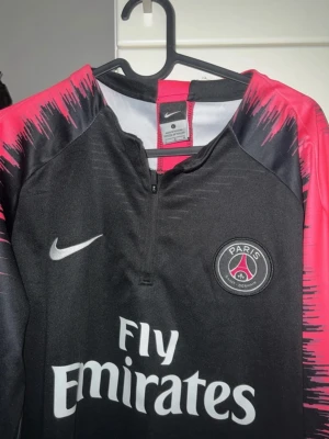Svart och röd PSG Nike träningströja - Säljer en snygg svart och röd träningströja från Nike med Paris Saint-Germain-logga på bröstet och Fly Emirates-tryck. Tröjan har halv dragkedja och coolt mönster på ärmarna. Perfekt för dig som gillar fotboll och vill ha något stilrent på träningen.