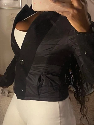 Svart quiltad kavaj med knappar - Snygg svart kavaj med quiltade partier och två stora knappar framtill. Jackan har en figurnära passform och är tillverkad i ett glansigt syntetmaterial. Långärmad och med markerad krage, perfekt för dig som gillar stilrena och moderna plagg.