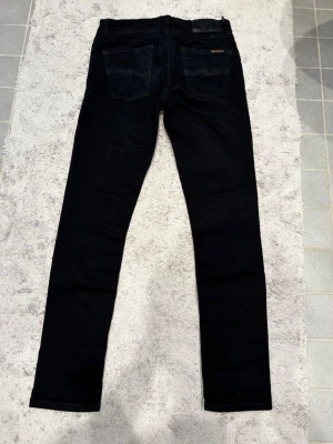 Svarta tight terry  jeans från Nudie Jeans - Säljer ett par svarta skinny jeans från Nudie Jeans i storlek W29 L28. Jeansen har klassisk femficksdesign, svarta sömmar och Nudie-logga på bakfickan. Materialet är mjukt och stretchigt för extra komfort.