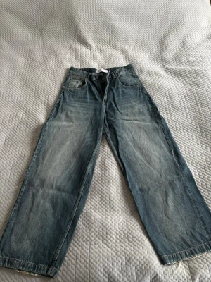Wide jeans från Bershka, blå - Säljer ett par blå jeans från Bershka i storlek 38 med breda ben och klassisk femficksdesign. Jeansen har dragkedja och knapp framtill, samt slitna detaljer vid benslut. Materialet är robust denim i bomull och passformen är loose.