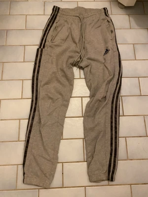 Grå mjukisbyxor från Adidas - Säljer ett par grå mjukisbyxor från Adidas med svarta ränder längs sidorna och logga på vänster ben. Byxorna har snörning i midjan och är gjorda i mjukt bomullsmaterial. Perfekt för chill eller träning, med normal passform och praktiska fickor.
