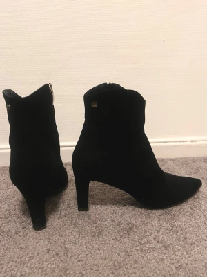 Svarta stövletter med klack och spetsig tå - Säljer ett par svarta stövletter med snygg spetsig tå och klack. Skorna är gjorda i mocka och har en dragkedja på sidan samt små metallknappar som detalj. Perfekta för dig som gillar stilrena och trendiga boots.