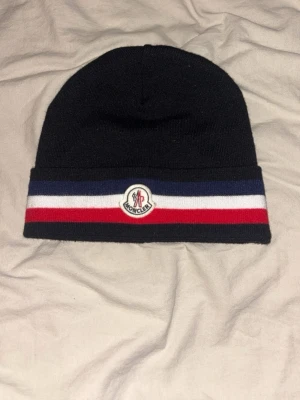 Moncler mössa med ränder - Helt splitter ny snygg svart Moncler mössa. Mössan har Moncler-logga framtill och är stickad i mjukt material som håller dig varm. Perfekt för dig som vill ha en stilren accessoar med lite lyxig känsla.
