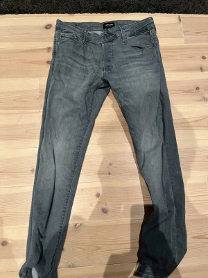 Grå jeans från Jack & Jones, storlek 31/30 - Snygga grå jeans från Jack & Jones med klassisk femficksdesign och smal passform. Jeansen har en diskret tvättad look och silverfärgad knapp framtill. Perfekt för dig som gillar stilrena och enkla jeans med modern känsla.