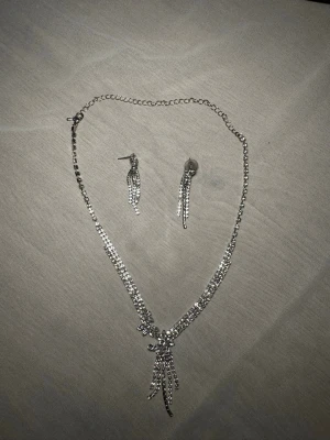 Glittrigt smyckeset i silverfärg - Säljer ett glittrigt smyckeset med halsband, armband och örhängen. Alla delar är dekorerade med gnistrande stenar och har en silverfärgad finish. Halsbandet har en hängande detalj framtill, örhängena har flera smala kedjor och armbandet har en stor sten i mitten.