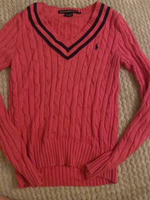 Rosa kabelstickad tröja Ralph Lauren - Säljer en rosa kabelstickad tröja från Ralph Lauren Sport i storlek M. Tröjan har v-ringad hals med mörkblå och rosa ränder, långärmad och med broderad logga på bröstet. Tillverkad i 100% bomull, perfekt för dig som gillar klassisk preppy stil.