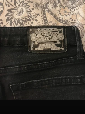 Svarta Crocker jeans bootcut - Säljer ett par svarta jeans från Crocker med klassisk femficksdesign och bootcut-ben. Jeansen har en snygg mörk tvätt och är tillverkade i stretchig denim som sitter skönt. Passar dig som gillar en tidlös look med lite utsvängda ben.