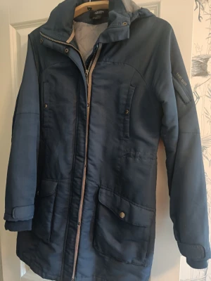 Blå parkajacka från Fresh Spirit - Snygg blå parkajacka från Fresh Spirit designad av Ellos. Jackan har huva, dragkedja framtill, tryckknappar och stora fickor med lock. Insidan är quiltad för extra värme och jackan har en stilren look som passar perfekt till kyliga dagar.