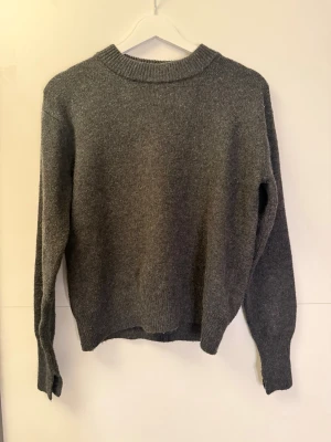 Stickad tröja från HM - Mysig mörkgrå stickad tröja med rund hals och långa ärmar. Tröjan har ribbade muddar vid hals, ärmslut och nederkant. Perfekt för kyliga dagar och enkel att matcha med jeans eller kjol.