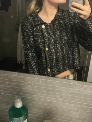 Svartvit croppad kofta med guldknappar - Säljer en croppad kofta i svart och vitt med snyggt rutigt mönster. Koftan har stora guldiga knappar framtill och en klassisk krage. Materialet är stickat och känns mjukt och lite tjockare, perfekt för kyligare dagar. Passformen är loose och ärmarna är långa.