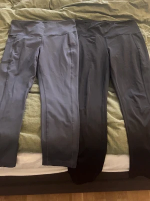 Svarta och mörkgrå leggings med fickor - Två par leggings i svart och mörkgrått, båda med praktiska sidofickor. De har hög midja och är tillverkade i ett stretchigt syntetmaterial som sitter tight mot kroppen. Perfekta för träning eller chill. Snygga sömmar och enkel design.