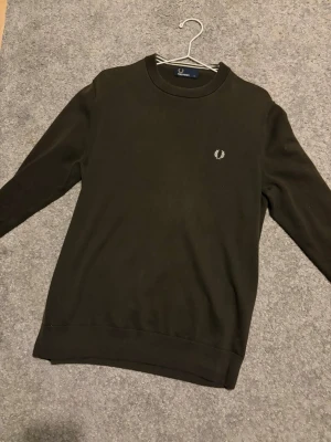 Mörkgrön stickad tröja Fred Perry - Snygg mörkgrön stickad tröja från Fred Perry i storlek M. Tröjan har rund hals, långa ärmar och ett broderat vitt Fred Perry-logo på bröstet. Perfekt för dig som gillar klassisk stil med en modern touch.