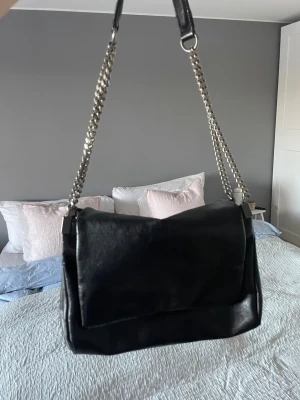 Svart axelväska med kedjerem Zara - Säljer en svart axelväska i skinn med bred lockstängning och snygg kedjerem i silverfärg.  Perfekt storlek för att få plats med allt du behöver under dagen. Från Zara 