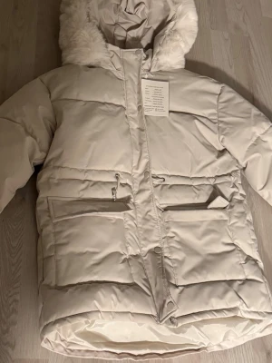 Beige pufferjacka  - Säljer en beige pufferjacka från Bamulan med stor huva och fuskpälsdetaljer. Jackan har dragkedja och tryckknappar framtill, samt två stora fickor med lock och dragkedja. Perfekt för kalla dagar och riktigt snygg till vinterlooken.