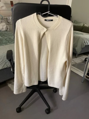 Vit/gul kofta från Gina Tricot - Säljer en stilren vit kofta från Gina Tricot med rund hals och en knapp framtill. Koftan har långärmad design och är tillverkad i ett mjukt stickat material som känns skönt mot huden. Perfekt att slänga över en topp när det är lite kyligt ute.