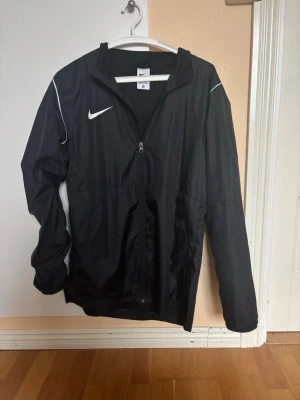 Svart nike Regnjacka - Säljer min Nike Regnjacka då jag har köpt en ny. Jackan är i väldigt bra skick och har inte använt den så mycket. Den är perfekt för dig som idrottar ute i regniga väder men passar även dig som vill använda den i vardagslivet.