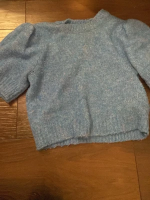 Blå stickad tröja från Vero Moda - Mysig blå stickad tröja från Vero Moda i storlek XS. Tröjan har korta puffärmar och en ribbad hals samt nederkant. Perfekt för dig som gillar en enkel men ändå trendig look med lite volym på axlarna.