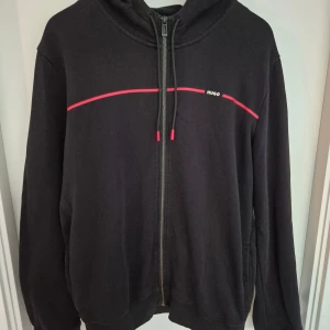 Svart hoodie från Hugo med röd detalj - Svart hoodie från Hugo med dragkedja och huva. Framsidan har en röd linje och diskret Hugo-logga på bröstet. Tröjan har långa ärmar, snörning i huvan och är tillverkad i mjuk bomull. Perfekt för en chill och stilren look.