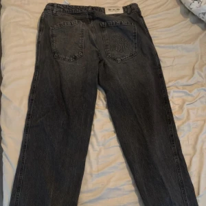 Svarta jeans från Black Squad - Säljer ett par svarta jeans från Black Squad med broderad detalj på bakfickan. Jeansen har klassisk femficksdesign, raka ben och dragkedja framtill. Materialet är jeans och färgen är tvättad svart med en lite sliten look. Perfekta för en avslappnad streetstil.