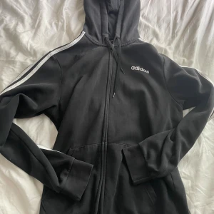 Svart Adidas hoodie med dragkedja - Svart hoodie från Adidas med vit logga på bröstet och klassiska vita ränder längs ärmarna. Tröjan har huva med snörning, dragkedja framtill och två fickor. Perfekt för chill eller träning, riktigt skön och stilren design.