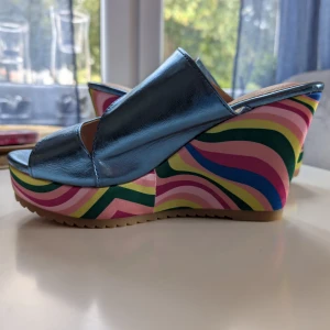 Blå metallic platåsandaler med mönster - Unika sandaler med blå metallic ovandel och öppen tå. Sulan är hög platå med färgglatt vågigt mönster i rosa, gult, grönt och blått. Skorna har breda remmar och plattformsdesign som ger extra höjd och statement-look. Perfekta för dig som vill sticka ut.                                       Helt oanvända!             Det är storlek 40 men dem passar mig strl.38.