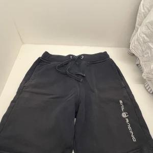 Svarta Sail Racing shorts med snörning - Svarta shorts från Sail Racing med vit logga längs benet. Mjukt material, troligen bomull eller polyester, och bekväm midja med snörning. Perfekta för chill dagar eller sport. Klassisk loose passform och praktiska fickor.