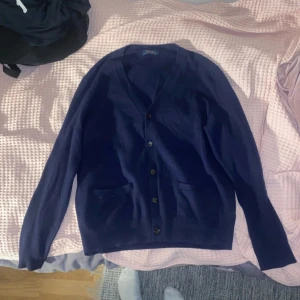 Marinblå cardigan från Polo Ralph Lauren - Snygg marinblå kofta från Polo Ralph Lauren med klassisk v-ringning och knappar framtill. Tröjan har två fickor och bruna mockadetaljer på armbågarna. Perfekt för dig som gillar stilrena och tidlösa plagg. Passar dig som vill ha en chill men ändå uppklädd look.