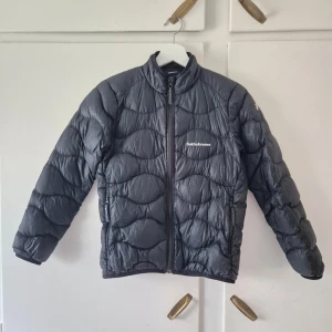 Svart lättviktsjacka Peak Performance - Svart lättviktsjacka från Peak Performance med vågformat quiltat mönster och dragkedja framtill. Jackan har hög krage och två sidofickor med dragkedja. Perfekt för kyliga dagar och enkel att matcha med olika outfits.