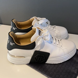 Philipp Plein vita sneakers med logga - Snygga vita sneakers från Philipp Plein med svart hälparti och tydlig logga med dödskalle. Skorna har vita snören, platt sula med silverdetalj och svart PP-märke på sidan. Tillverkade i skinn med beige innersida för extra komfort.