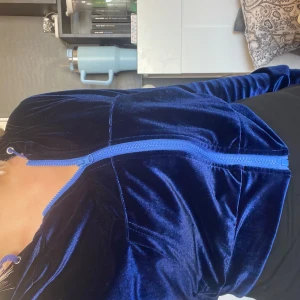 Juicy Couture blå sammetshoodie - Säljer en ikonisk mörkblå hoodie i sammet från Juicy Couture med glittrande Juicy-text i strass på ryggen. Tröjan har dragkedja framtill, huva och långa ärmar. Perfekt för dig som vill ha en lyxig och trendig look.