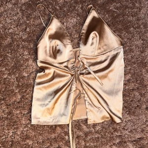 Champagnefärgad satin linnetopp - Snygg champagnefärgad linnetopp i satin med smala axelband och djup v-ringning. Toppen har snörning framtill som ger en cool detalj och croppad passform. Perfekt att styla med jeans eller kjol för en trendig look.