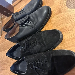 Svarta finskor från Gant och läderskor - Två par klassiska svarta finskor, ett par i mocka från Gant med rund tå och snörning, samt ett par i slätt svart skinn med snörning. Perfekta till kostym eller dressade outfits. Båda paren har platt sula och stilren design.