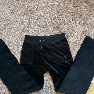 Svarta Juicy Couture  - Svarta mjukisbyxor i mjuk sammet med bred resår i midjan och snörning framtill. Byxorna har stora fickor fram och en diskret broderad logga på ena fickan. Perfekta för en chill och avslappnad stil.